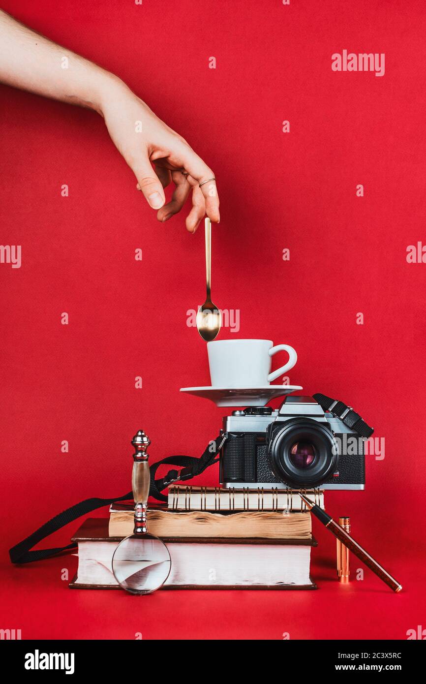 Balancing still life composition with books, vintage camera, cup of coffee and spoon toched by woman's hand on red background Stock Photo