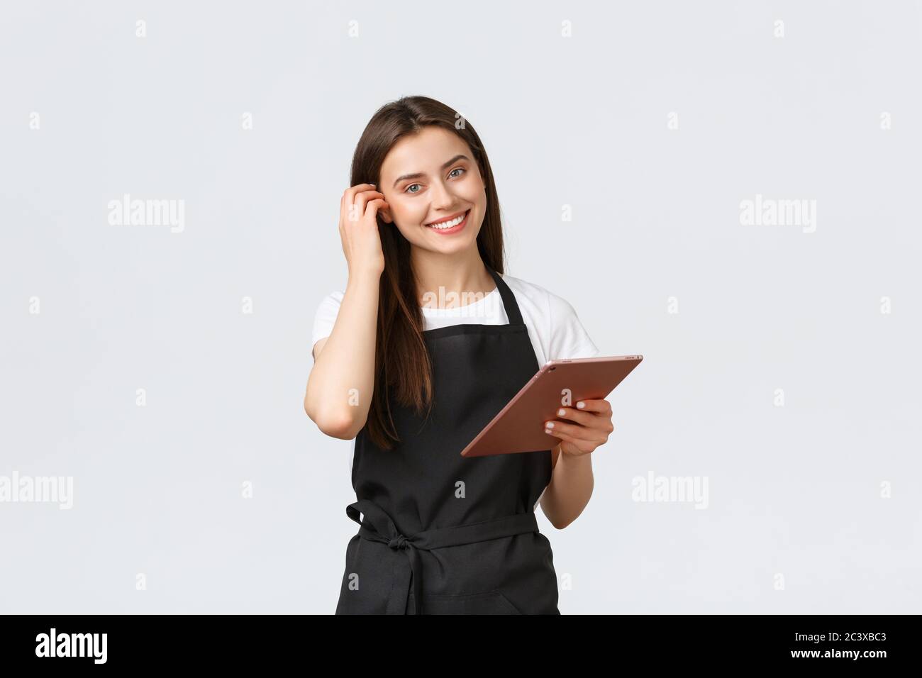 Grocery store employees, small business and coffee shops concept. Friendly cute female barista in black apron smiling at camera, using digital tablet Stock Photo