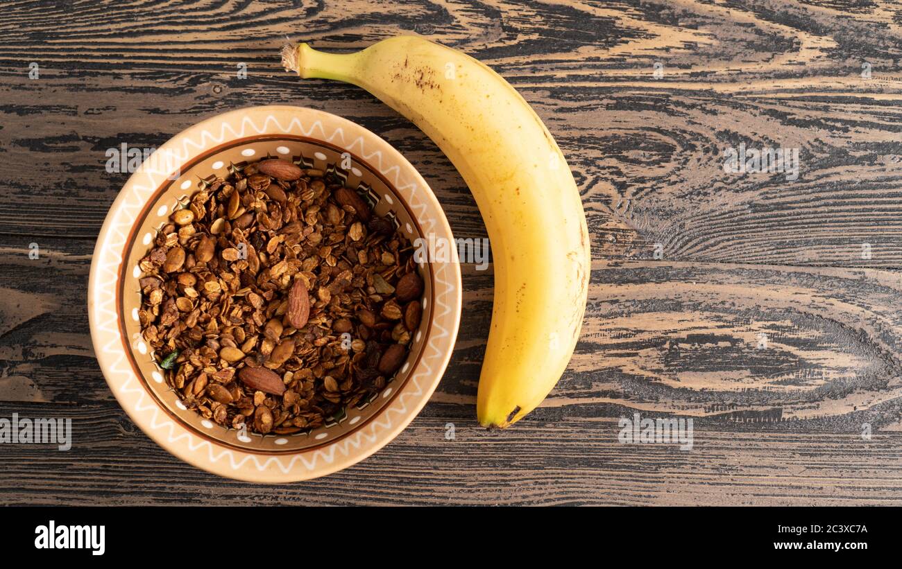 Homemade granola with nuts in a brown bowl and banana. Delicious and hearty breakfast. Rustic wooden table Stock Photo
