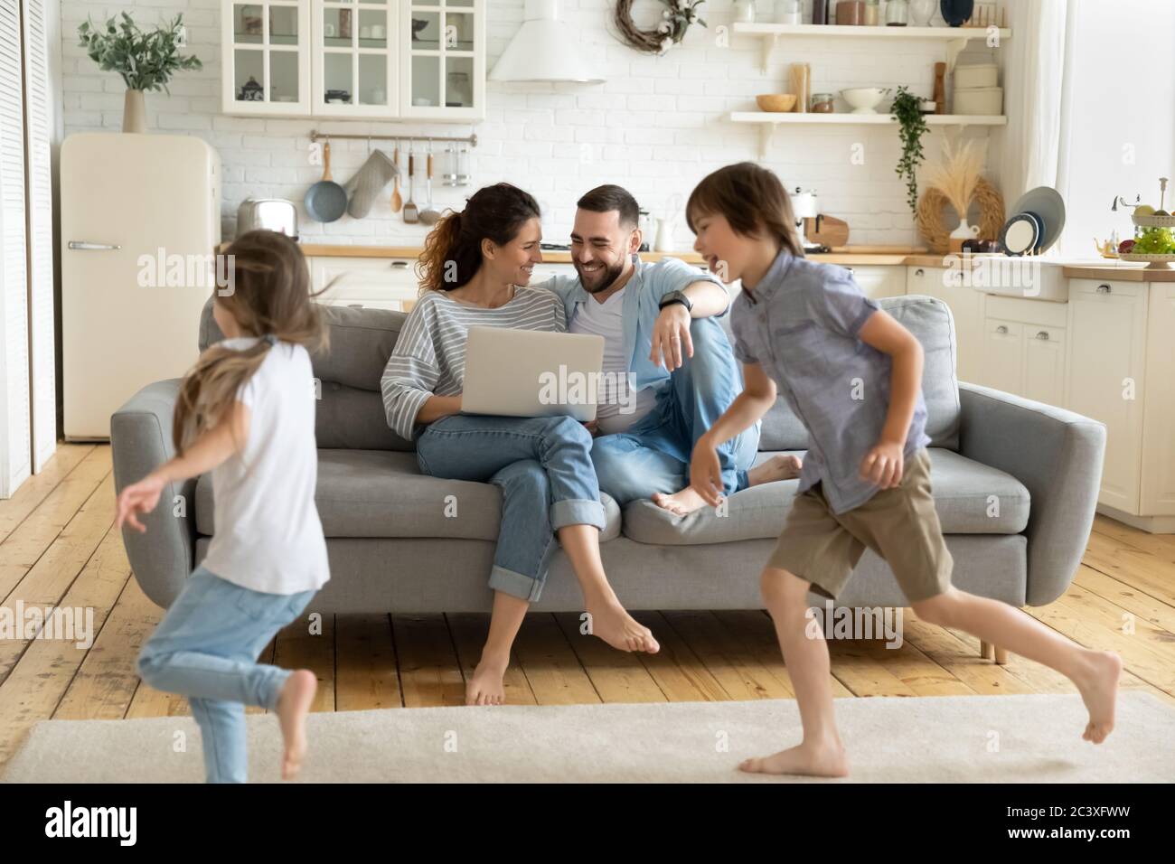 Happy young couple sitting at couch near running children. Stock Photo