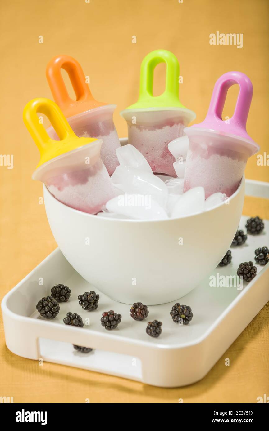Four homemade blackberry popsicles in a white bowl of ice, in a white tray with fresh blackberries, with a yellow background Stock Photo