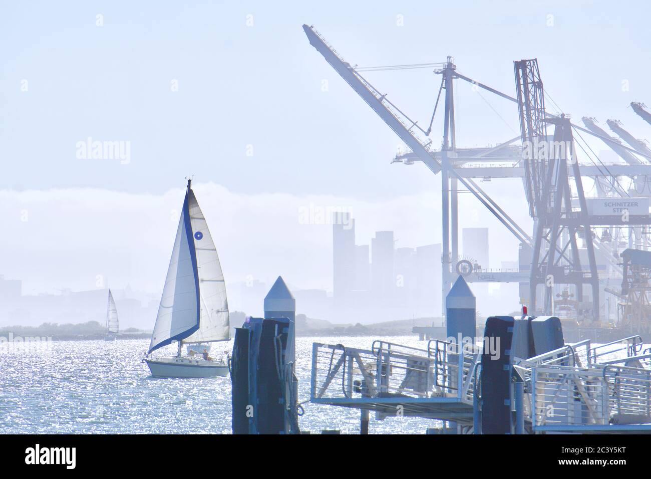 Sailboat entering Oakland Inner Harbor, at Jack London Square, with shipping cranes and docks. Bay Area tourism and recreation. Oakland, CA, USA. Stock Photo