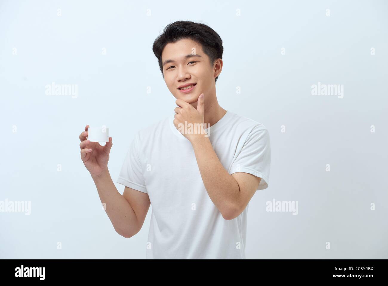 Young man touching his chin, holding face cream on hand, smiling
