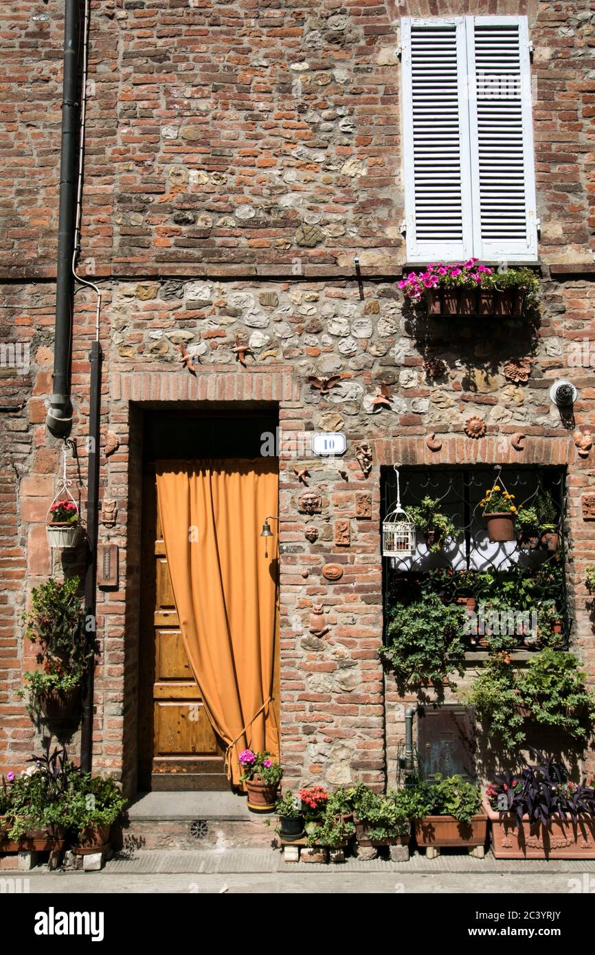 Detail of a typical facade of a house in a small Italian village with brick wall, a doorway with curtain and flowering plants in pots and at the windo Stock Photo