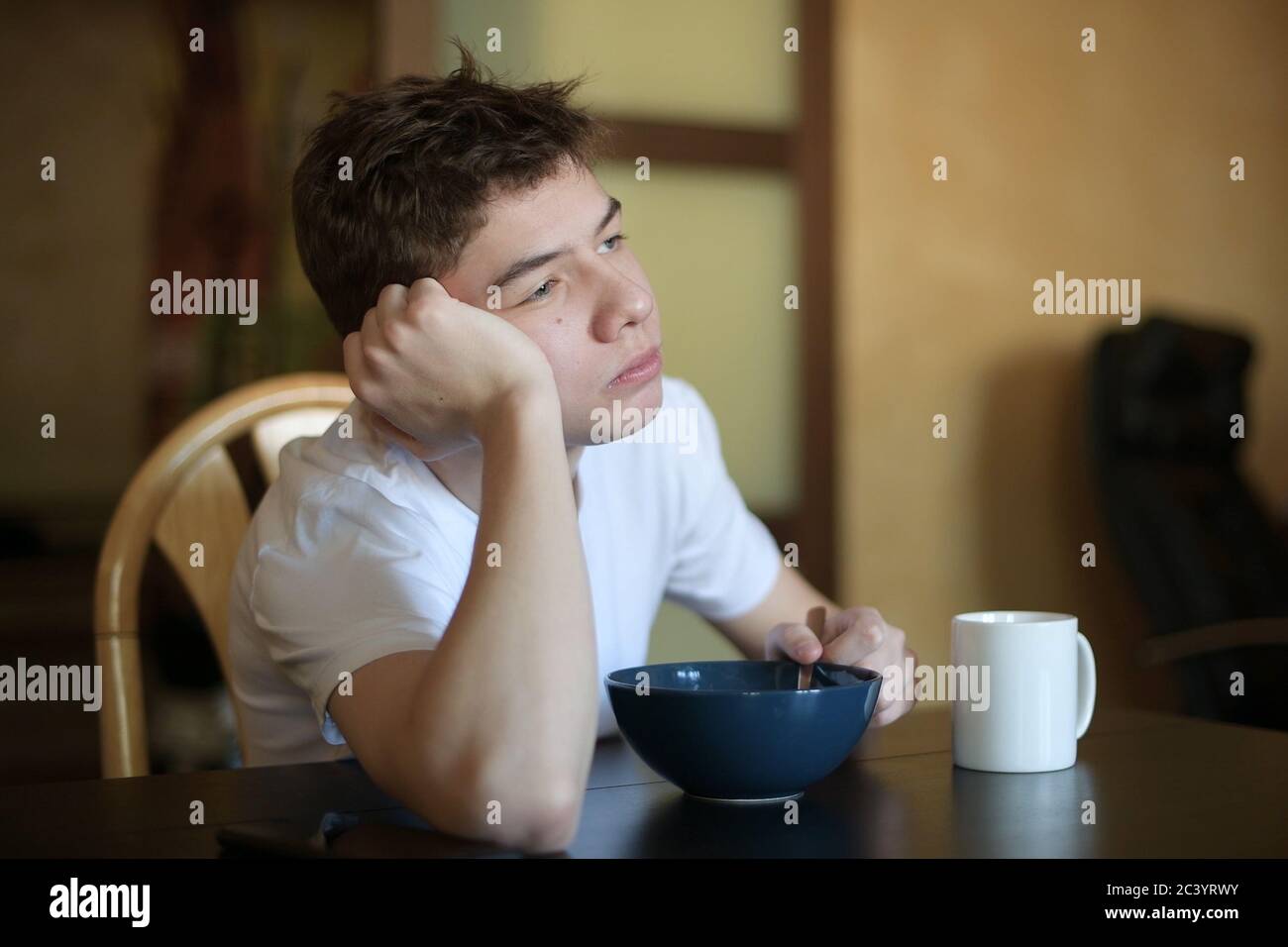 A teenager with a sleepy face idly looks out the window and chews rice porridge Stock Photo