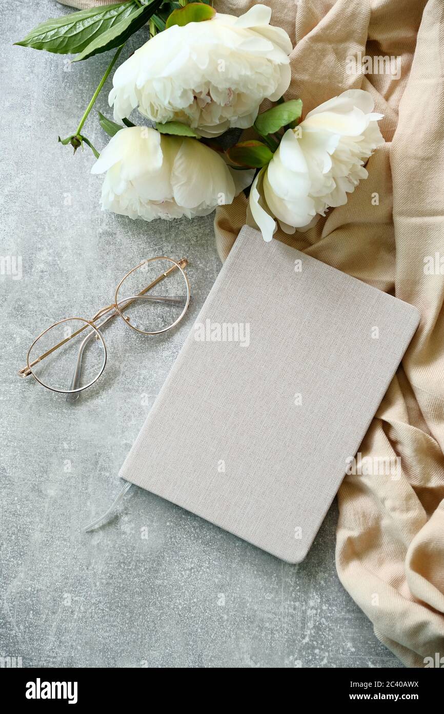 Female workspace with paper notebook, hipster glasses, beige cloth and peony flowers on stone table. Top view. Romantic flat lay style composition. Stock Photo