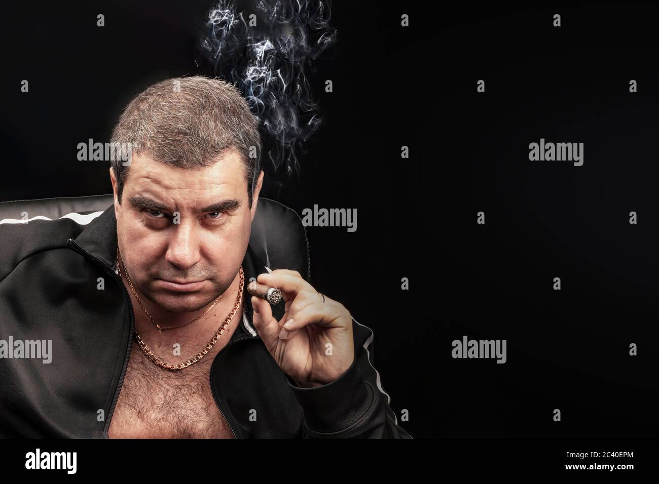 a stern adult man Smoking a cigar looks confidently at the camera. copy space on a black background. a crime boss, a mobster serving time in prison. Stock Photo