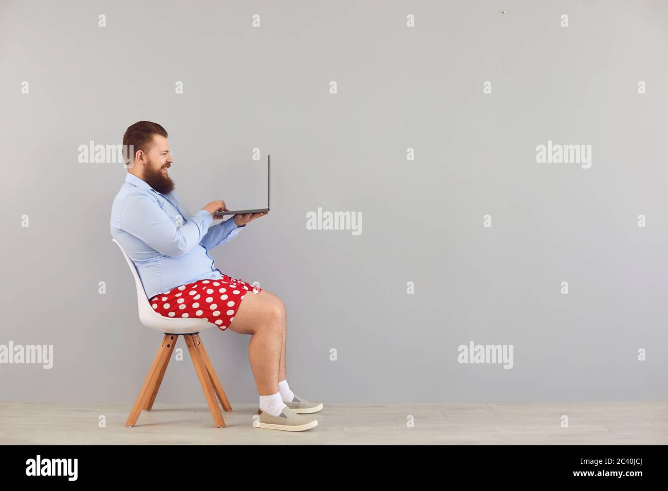 Funny fat man in a blue shirt and red shorts sitting on a chair working online using a laptop on a gray background. Stock Photo