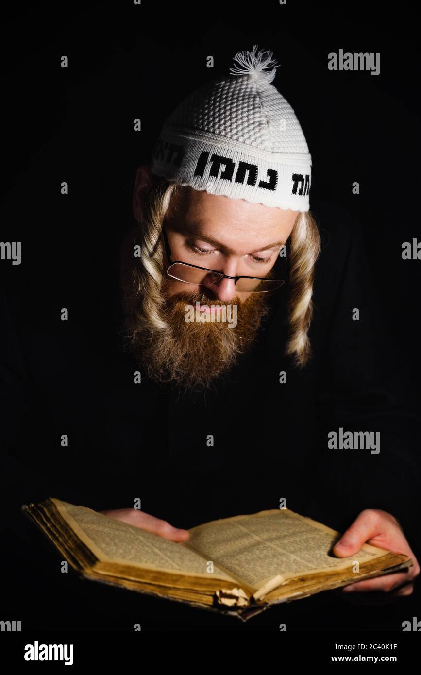Hasidic jew reading Torah. Religious orthodox jew with sidelocks and red beard in white bale praying in the dark. Low key photo. Vertical orientation Stock Photo