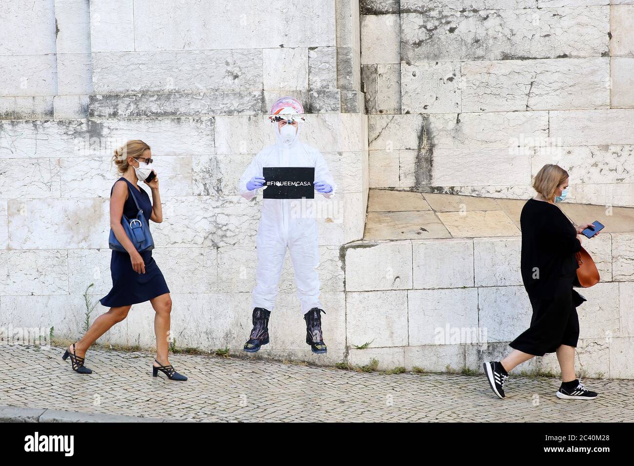 June 23, 2020, Lisbon, Portugal: People wearing face masks walk by a paper figure of a firefighter dressed in protective gear and holding a Ã”stay at homeÃ sign on a wall in Lisbon, Portugal, on June 23, 2020, during the COVID-19 Coronavirus pandemic. The Government applies today new mandatory confinement measures to the whole Lisbon metropolitan area, due to a significant increase in Covid-19 cases in recent weeks. Among the main restrictions is the return of the ban on gatherings with more than ten people, the strengthening of the supervision of shopping centres and the general closure of s Stock Photo