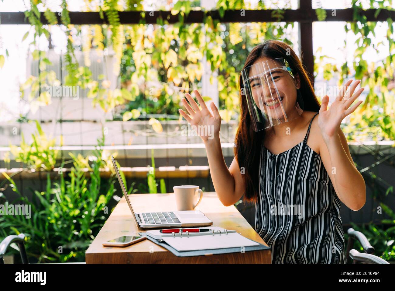 Smart young Asian woman smiling and waving hand while sitting at table ...