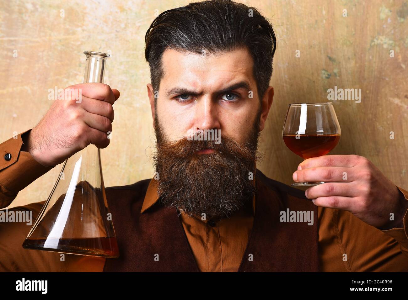 Man with beard holds alcoholic beverage on beige wall background. Service and restaurant drinks concept. Macho with serious face drinks brandy or whiskey. Guy with glass and bottle of cognac Stock Photo