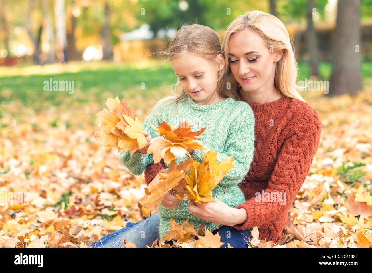 Happy young caucasian woman and little girl holding autumn yellow leaves sitting at the park Stock Photo