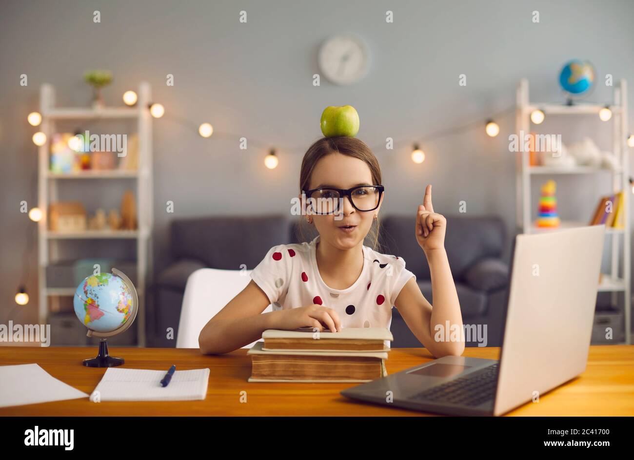 Adorable little girl with apple on her head and heap of books pointing up in front of laptop computer at home Stock Photo