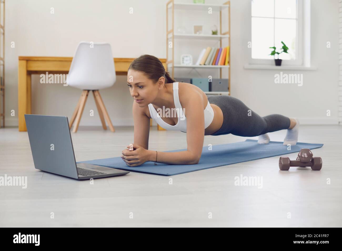 Young girl doing elbow plank in front of laptop computer at home. Fit woman exercising to online sports class indoors Stock Photo