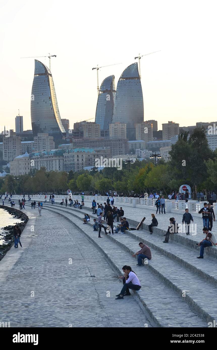 People walking and cycling along the Baku Boulevard, with the Caspian Sea on one side and city buildings on the other.