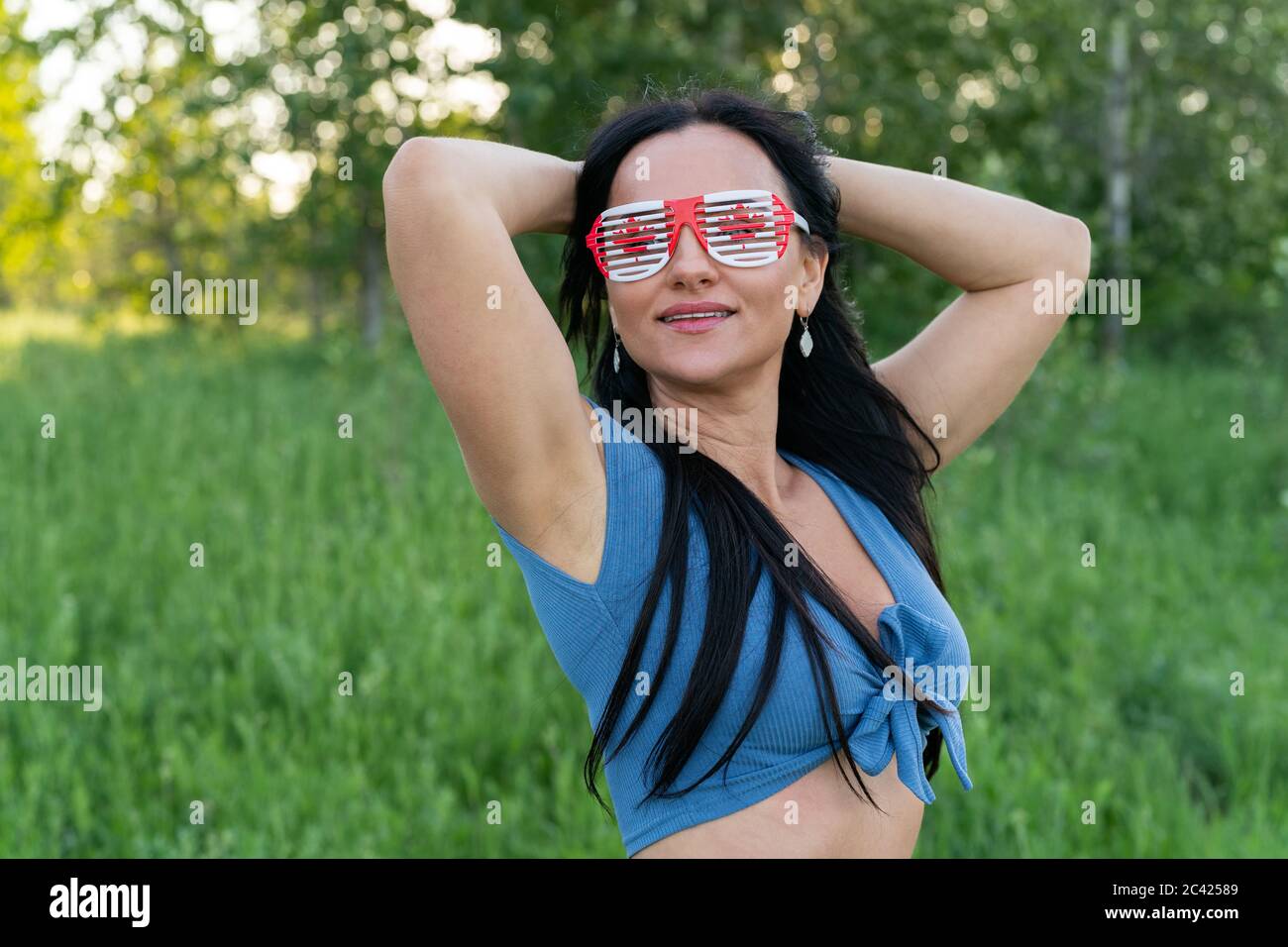 Happy Canada Day Celebration concept. Brunette woman with Canadian flag and themed sunglasses on nature background. Stock Photo
