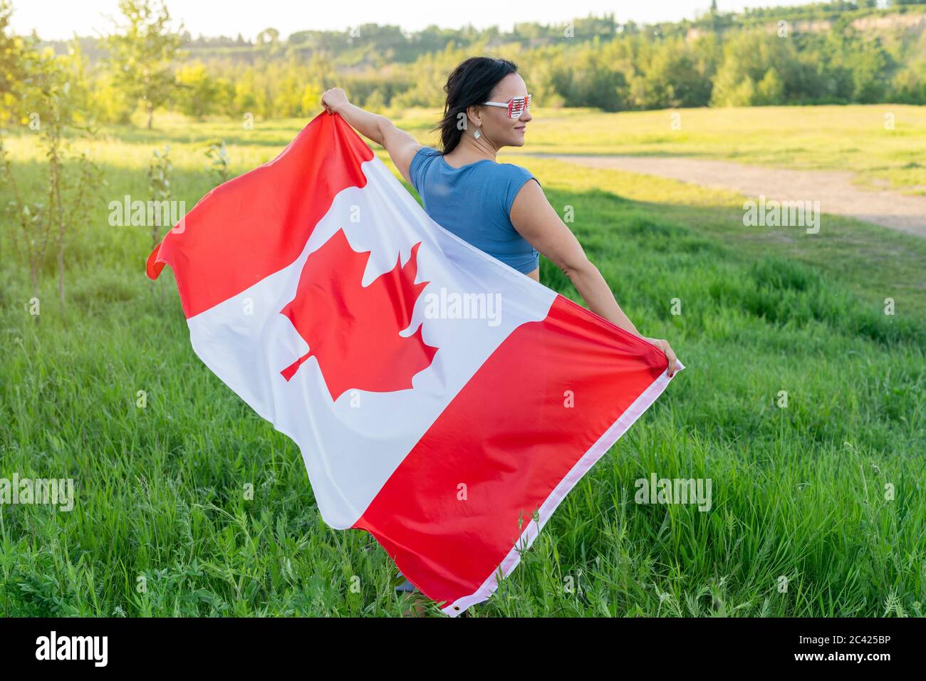 Happy Canada Day Celebration concept. Brunette woman with Canadian flag and themed sunglasses on nature background. Stock Photo