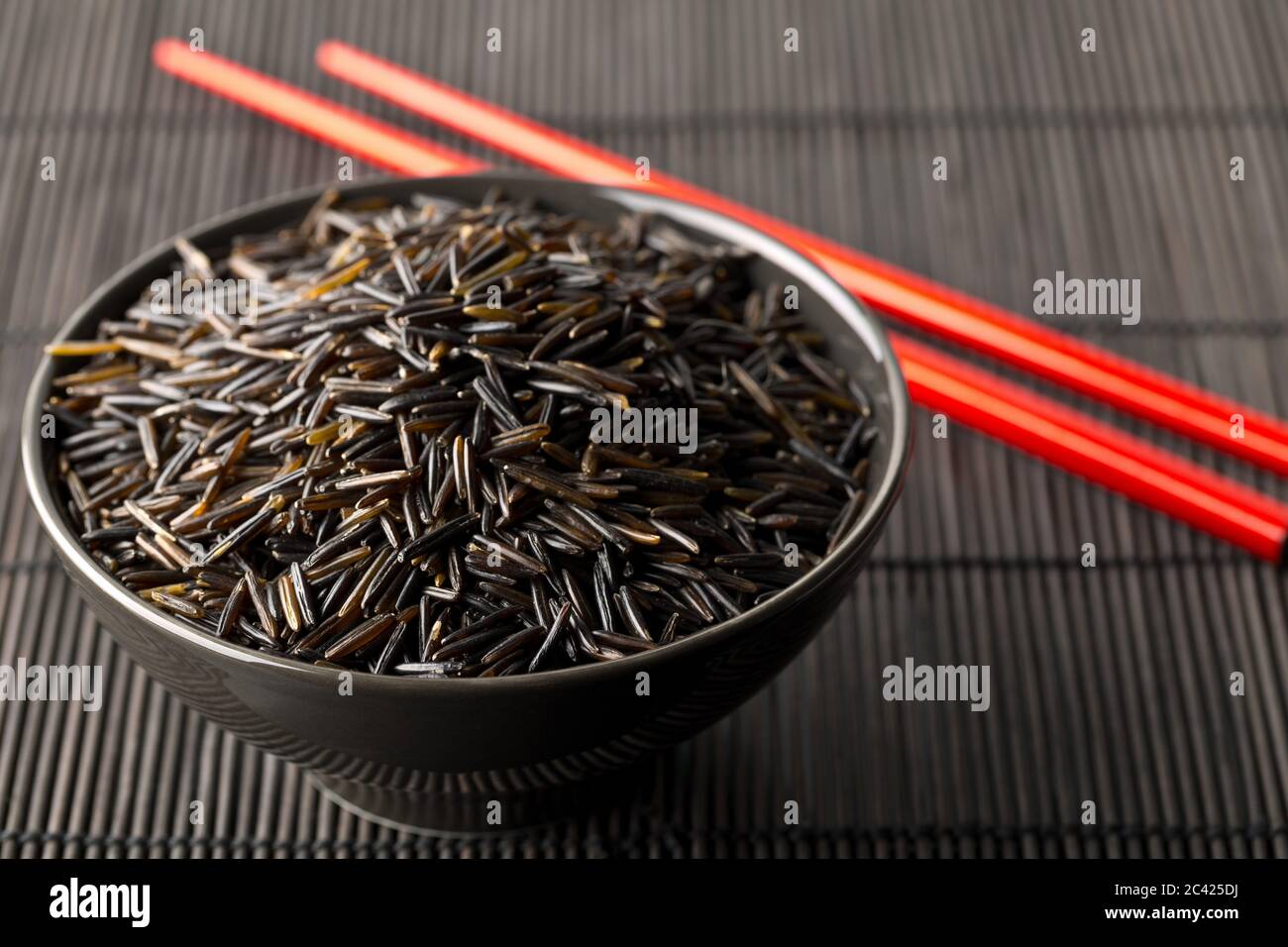 Heap of uncooked, raw, black wild rice grains in black bowl with red chopsticks on black bamboo mat background, selective focus Stock Photo
