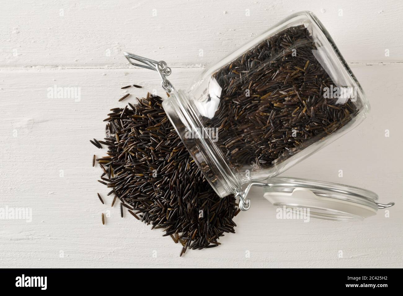 Heap of uncooked, raw, black wild rice grains in glass storage jar on white table background, selective focus, flat lay top view from above Stock Photo