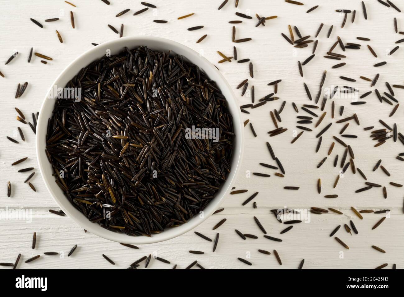 Heap of uncooked, raw, black wild rice grains in white bowl on white wooden table background, selective focus, flat lay top view from above Stock Photo