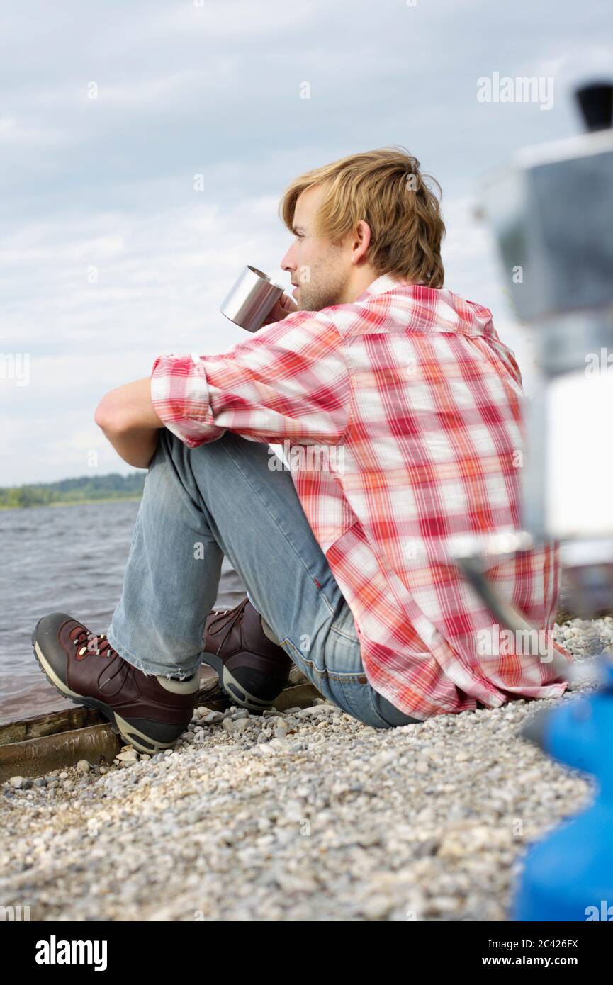 Young man sits by the water and drinks coffee Stock Photo