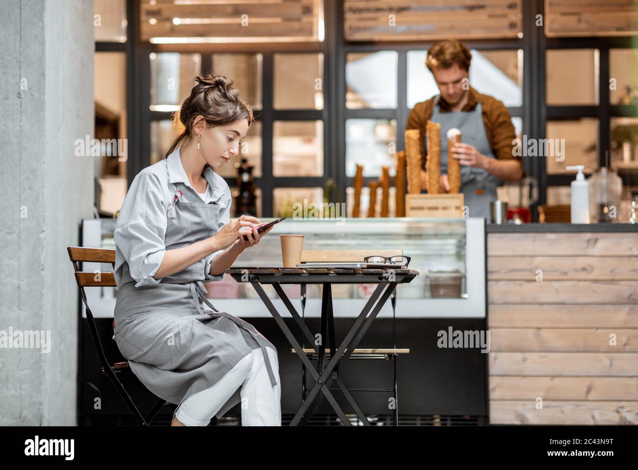 Working staff at the cafe or pastry shop, waiter sitting with phone and salesperson having some work at the shop counter Stock Photo