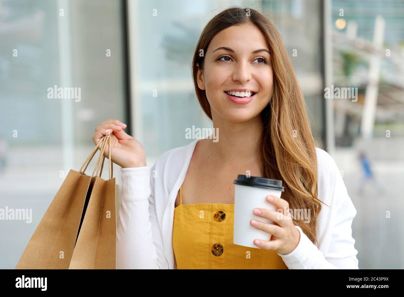 Positive young smiling woman looking aside with shopping bags and cup of cappuccino in her hands Stock Photo