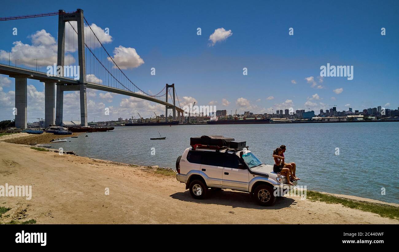 Mozambique, Katembe, Adult couple sitting on hood of 4x4 car admiring view of Maputo Bay with city and Maputo-Katembe Bridge in background Stock Photo