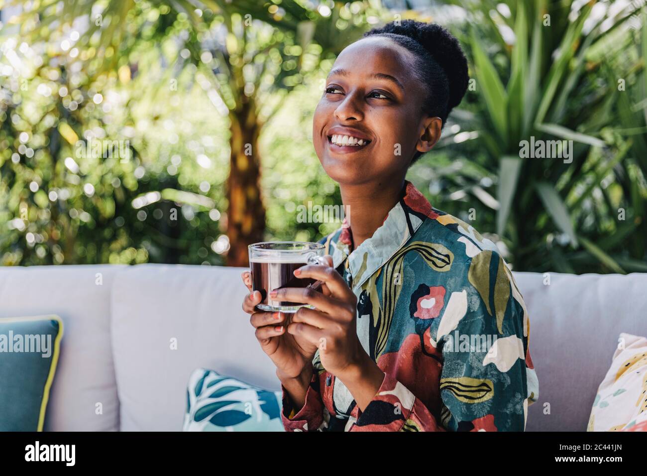 Portrait of happy young woman sitting on couch in garden drinking black coffee Stock Photo