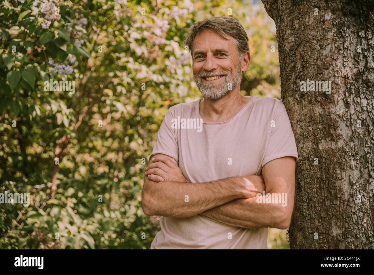 Smiling bearded man with arms crossed leaning on tree trunk at garden Stock Photo