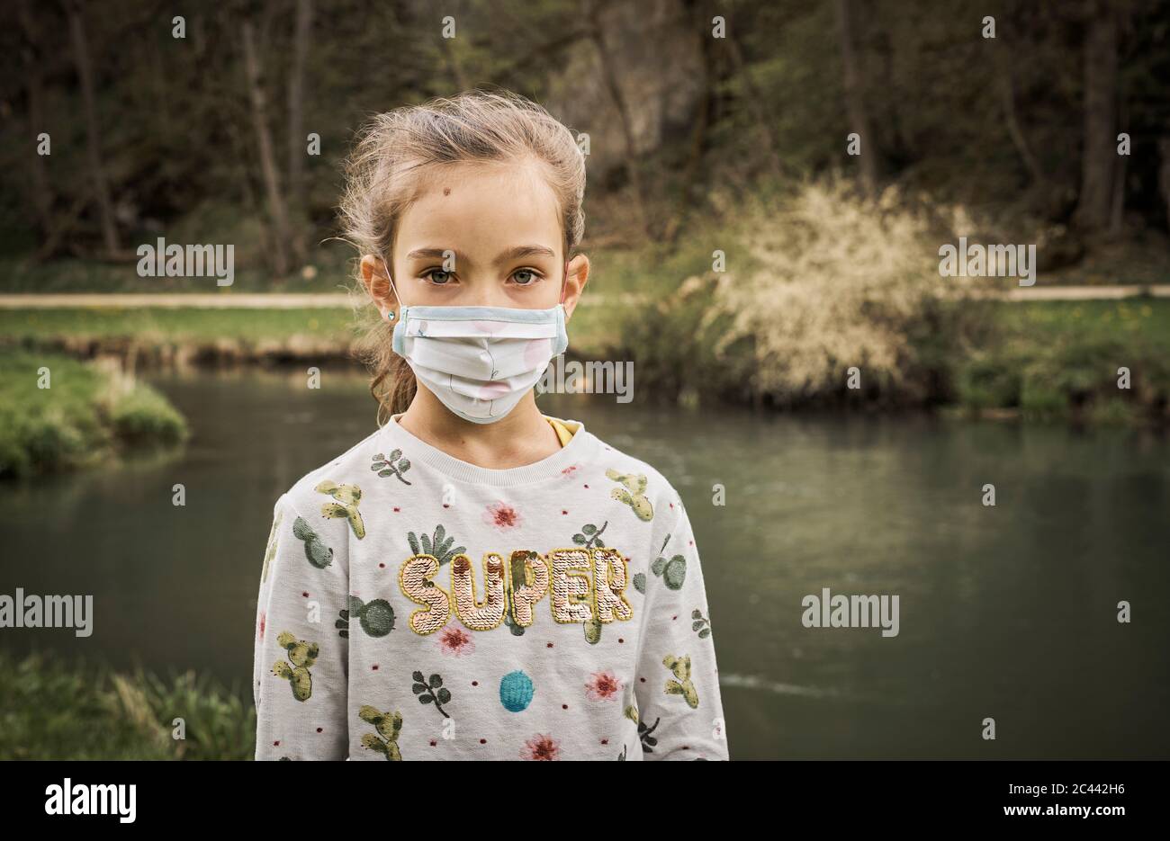 Portrait of sad girl standing with self-sewn face mask at riverbank in forest Stock Photo