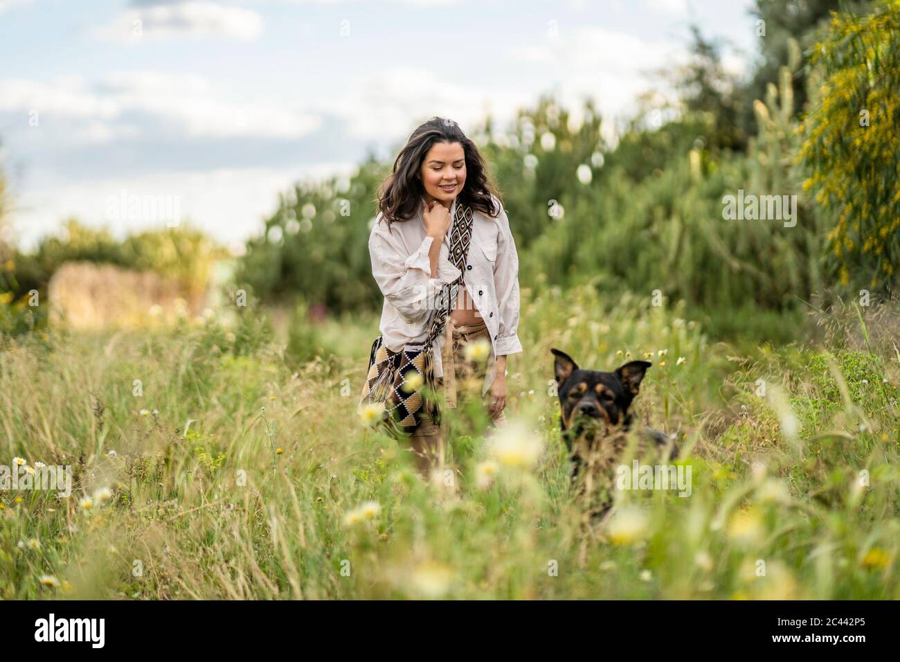Smiling beautiful young woman walking with dog on green plants against sky, Alicante, Alicante Province, Spain Stock Photo