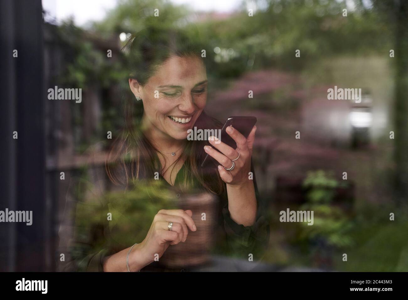 Smiling mid adult woman using mobile phone at home seen through window Stock Photo