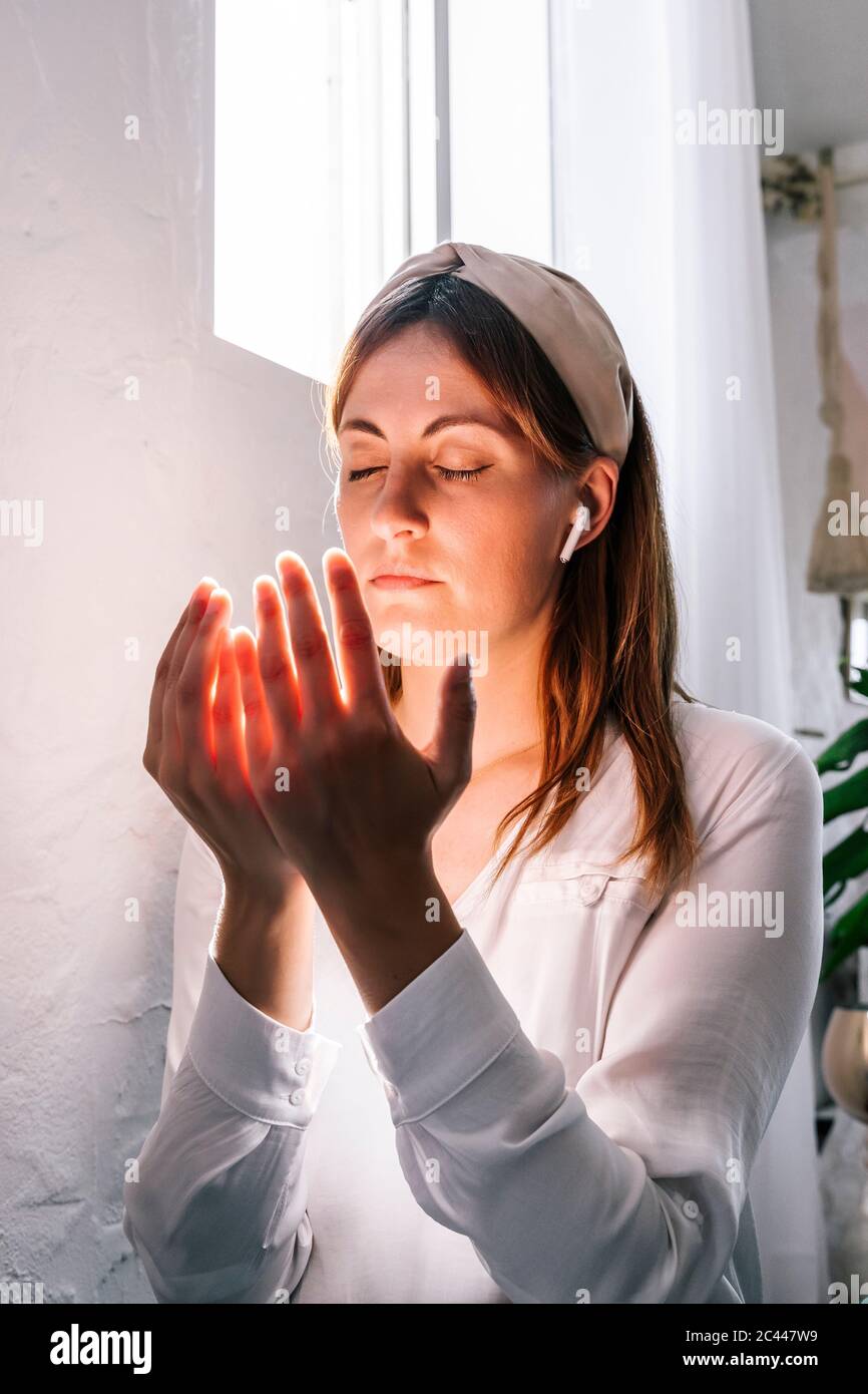 Beautiful woman praying with eyes closed by window at home Stock Photo