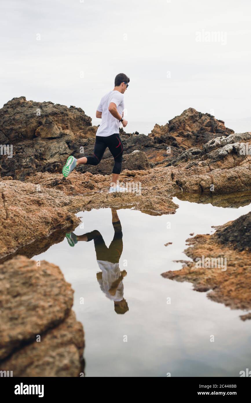 Full length of male trail runner on rocks while running by his reflection over water at coast, Ferrol, Galicia, Spain Stock Photo