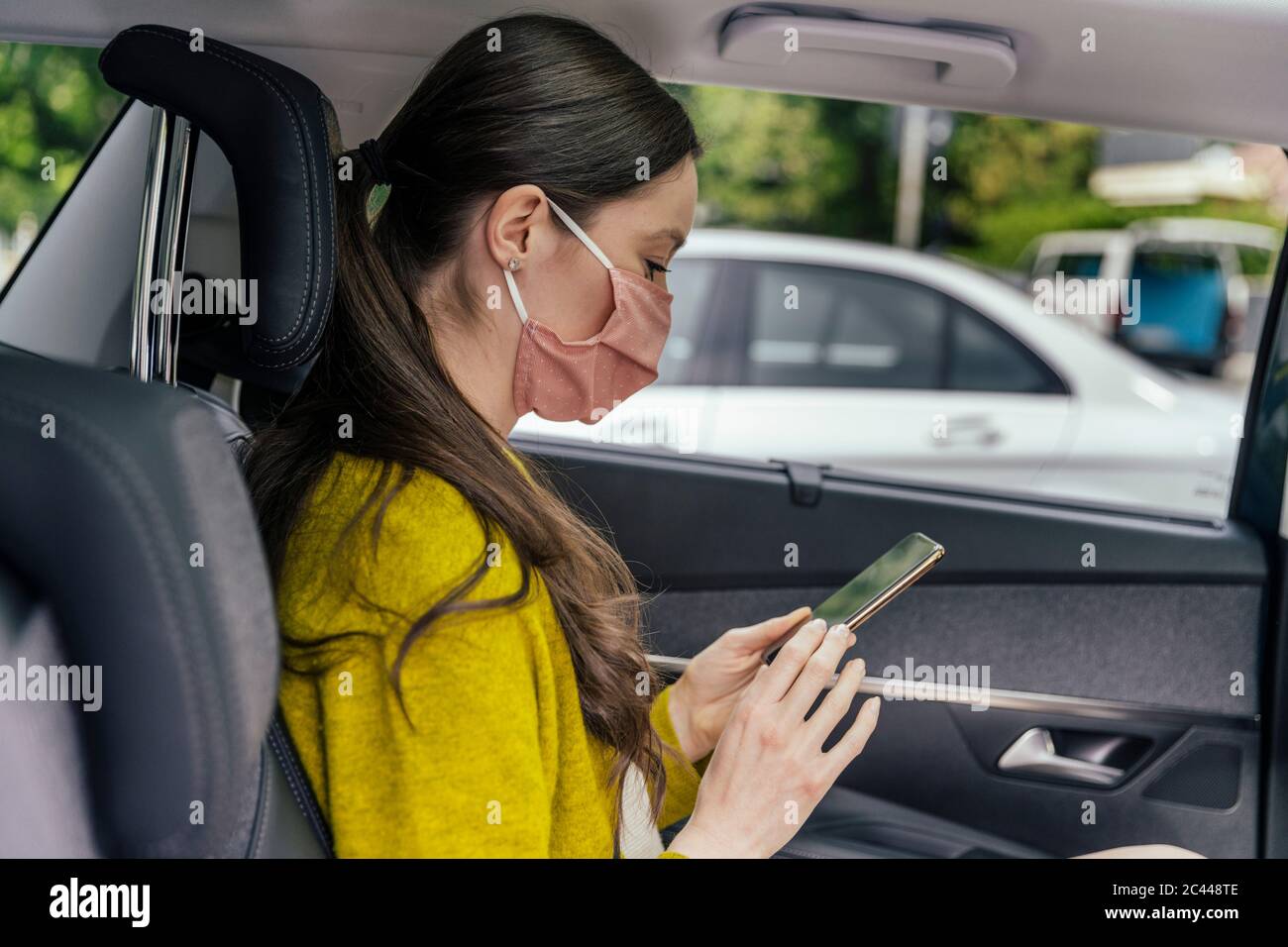 Young woman wearing protective mask sitting on back seat of car using cell phone Stock Photo