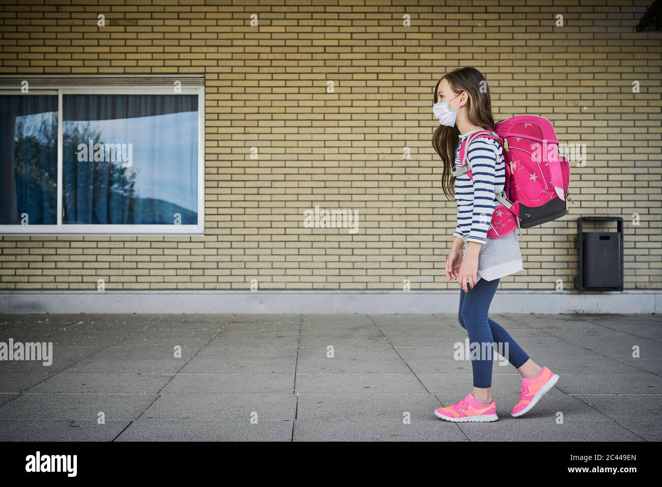 Girl wearing mask and schoolbag walking along building Stock Photo