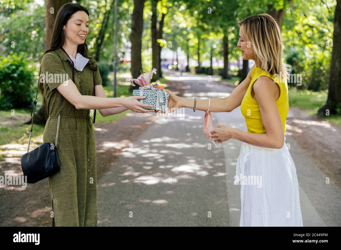Woman with taken off face mask handing over gift to friend in  nature Stock Photo