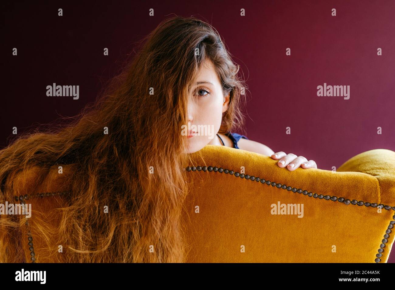 Close-up portrait of young woman with long brown hair leaning on golden chair against colored background Stock Photo