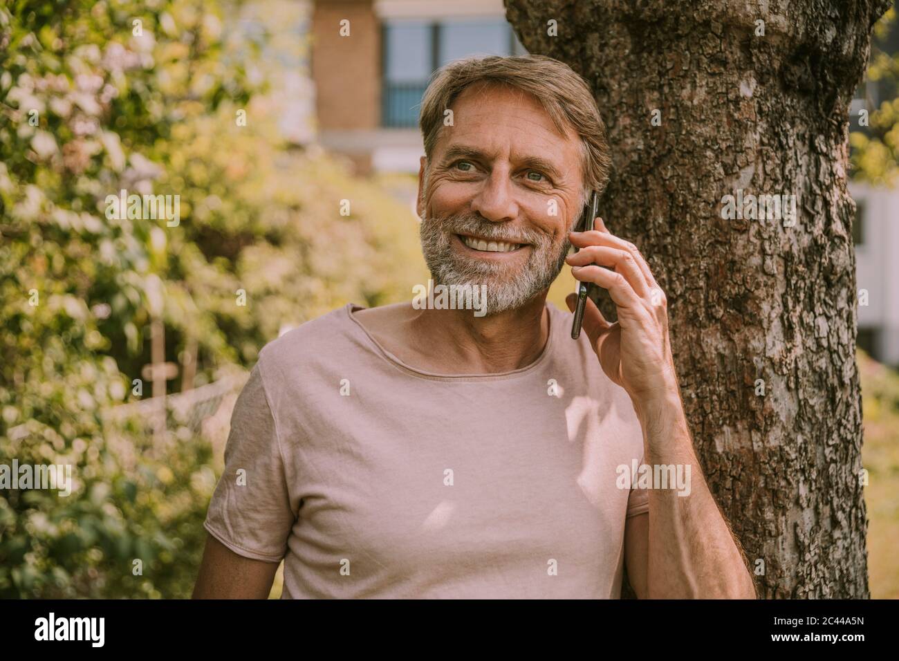 Smiling mature man looking away while talking on mobile phone against tree at garden Stock Photo