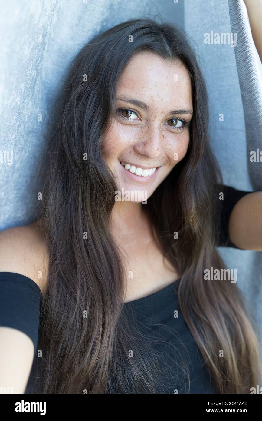 Close-up portrait of smiling young freckled woman against curtain at home Stock Photo