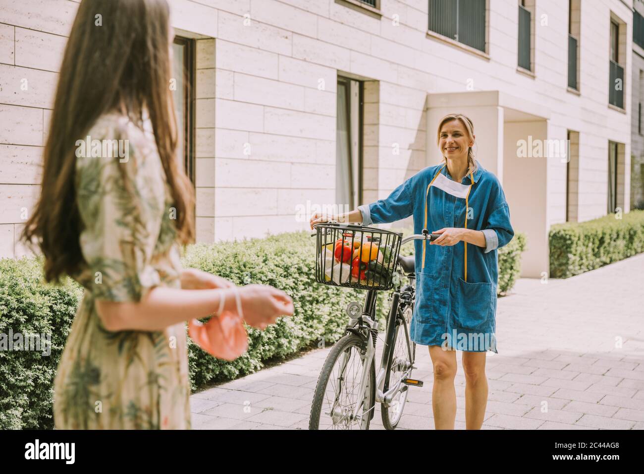 Two women with bicycle and face mask meeting in urban area Stock Photo