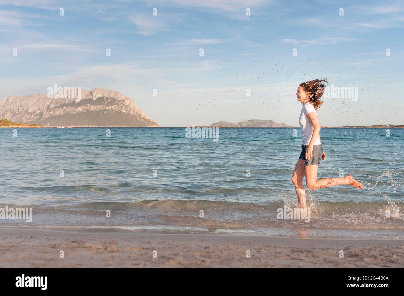 Happy woman running at seashore splashing with water, Sardinia, Italy Stock Photo