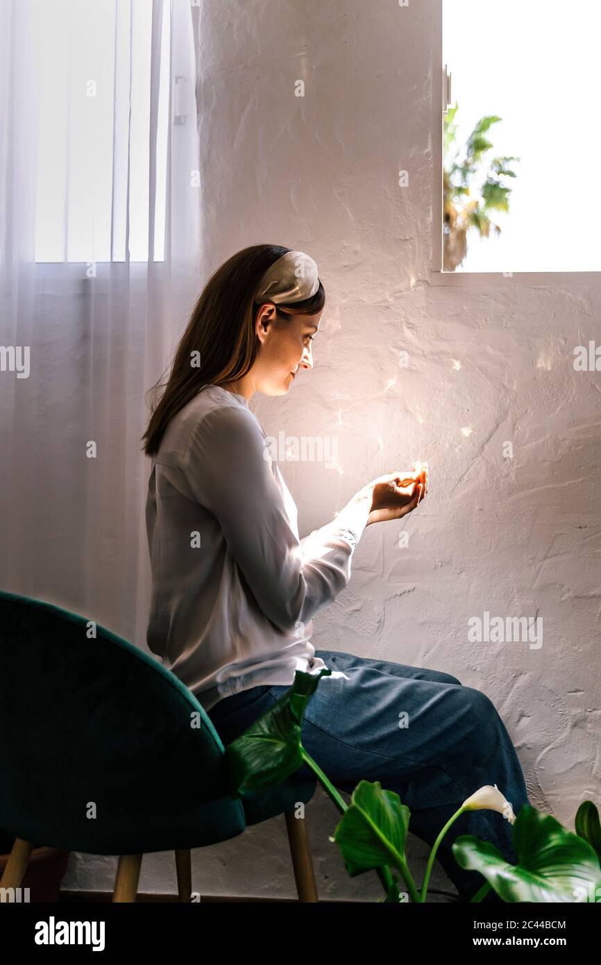 Woman sitting on chair while looking at sunlight on cupped hands in bedroom Stock Photo