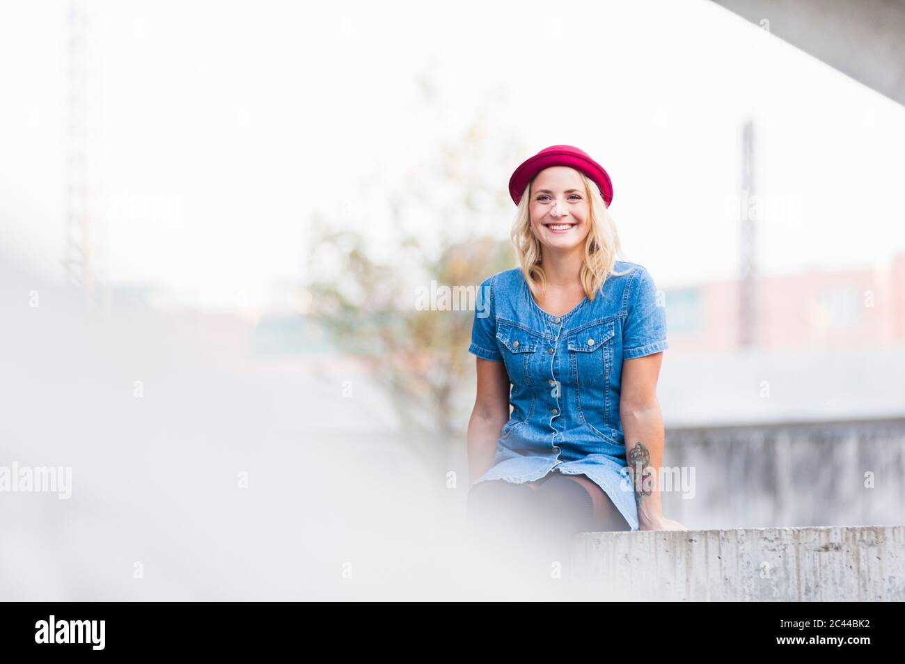 Portrait of happy young woman wearing denim dress sitting on a wall Stock Photo