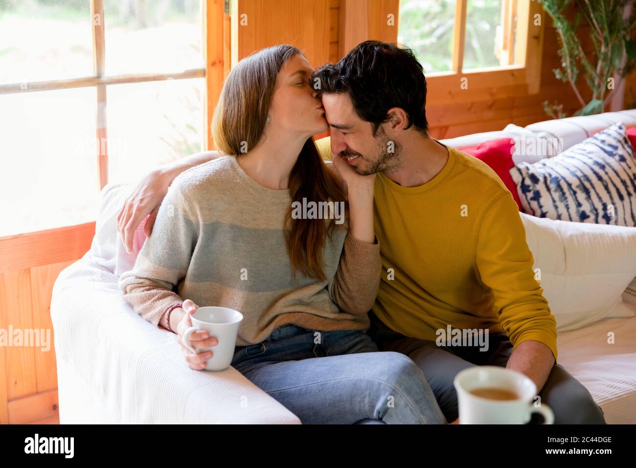 Romantic woman kissing on boyfriend's forehead while sitting over sofa in log cabin Stock Photo