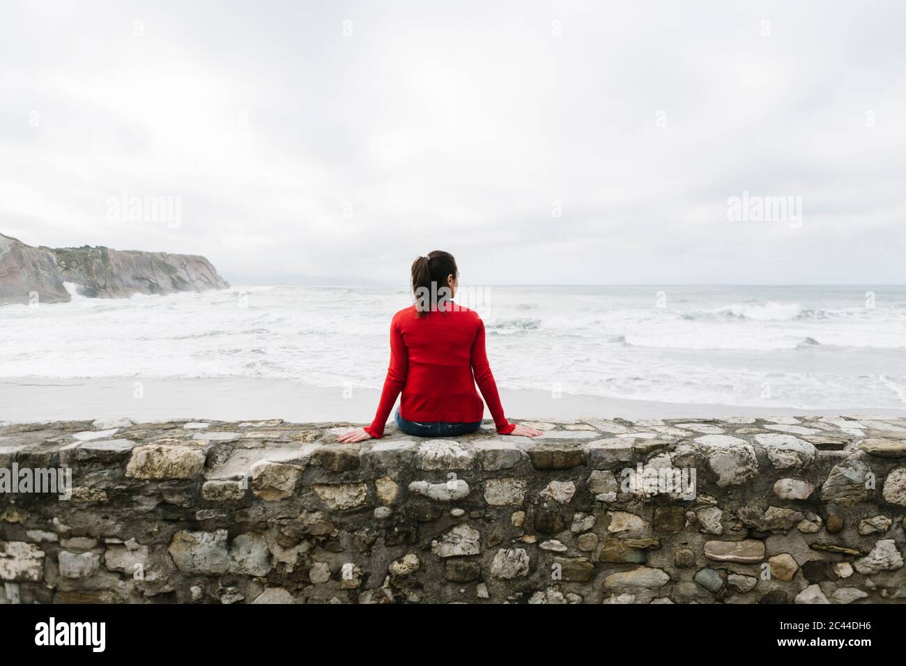 Rear view of relaxed female tourist sitting on retaining wall while looking at sea against sky, Itzurun, Zumaia, Spanish Basque Country, Spain Stock Photo