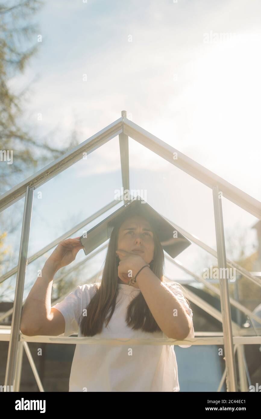 Pensive young woman covering head with laptop while standing in incomplete greenhouse against sky Stock Photo