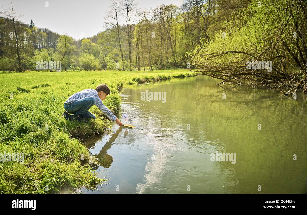Boy puts his self-made toy boat into river in forest on sunny day Stock Photo